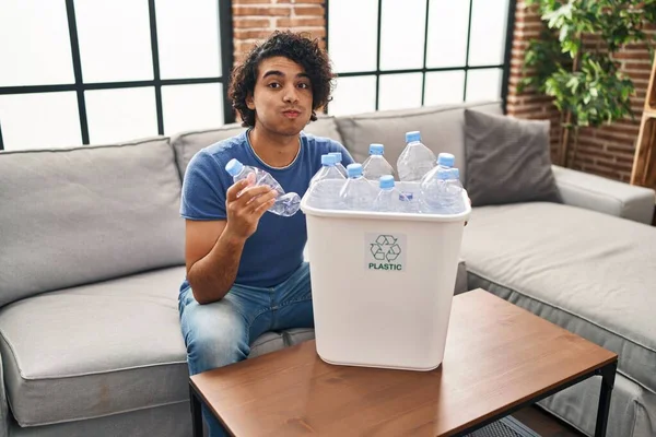 Hispanic man with curly hair holding recycling bin with plastic bottles at home puffing cheeks with funny face. mouth inflated with air, catching air.