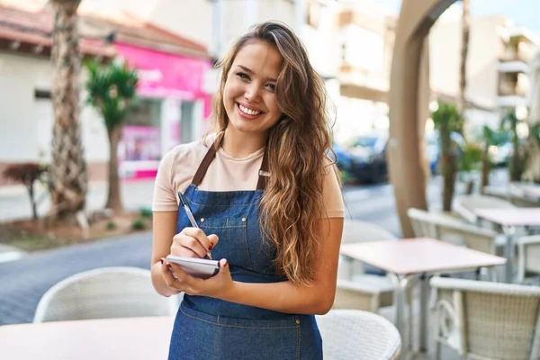 Young beautiful hispanic woman waitress smiling confident writing on notebook at coffee shop terrace