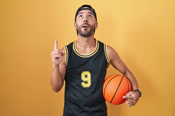 中年秃头男子在黄色的背景下拿着篮球 惊讶地抬起头 用手指和胳膊指着球 — 图库照片