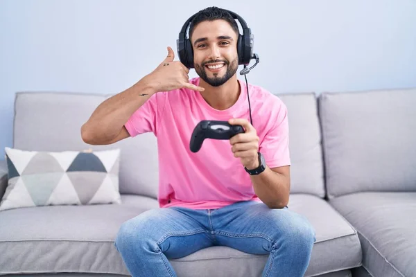 西班牙裔年轻人拿着控制器坐在沙发上玩电子游戏 微笑着用手和手指做着电话手势 就像在讲电话一样 交流概念 — 图库照片