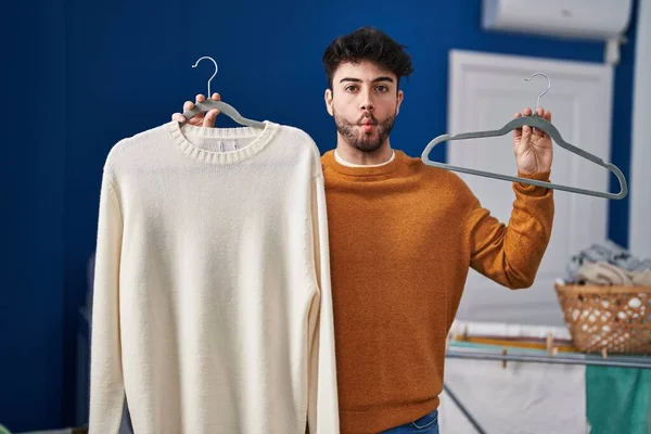 stock image Hispanic man with beard holding sweater on hanger at laundry room making fish face with mouth and squinting eyes, crazy and comical. 