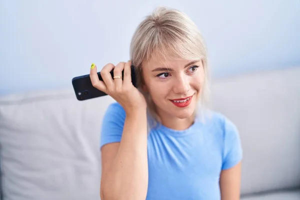 Młoda Blondynka Słuchanie Wiadomości Głosowej Przez Smartfona Siedzi Kanapie Domu — Zdjęcie stockowe
