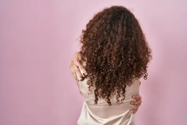 头发卷曲的西班牙裔女人站在粉红的背景上 高兴地拥抱自己 从后面积极地拥抱自己 自爱和自我照顾 — 图库照片