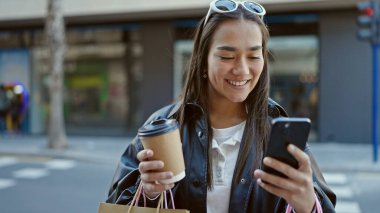 Genç ve güzel İspanyol kadın akıllı telefon kullanıyor. Elinde alışveriş poşetleri ve sokakta kahve tutuyor.