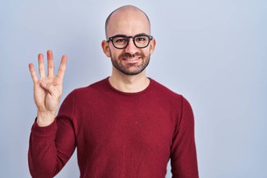 Beyaz arka planda duran sakallı genç kel bir adam gözlük takıyor ve dört numaralı parmaklarıyla işaret ederken kendinden emin ve mutlu bir şekilde gülümsüyor.. 