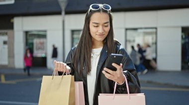 Genç, güzel, İspanyol bir kadın akıllı telefon kullanıyor. Elinde alışveriş torbaları var.