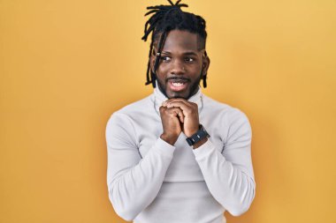 Sarı arka planda balıkçı yaka kazak giyen Afrikalı bir adam gergin ve heyecanlı bir şekilde çenesinin kenarına bakıyor. 