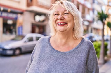 Orta yaşlı sarışın kadın sokakta kendinden emin bir şekilde gülümsüyor.
