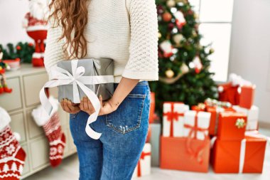 Young beautiful hispanic woman holding christmas gift on back at home