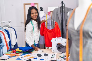 Young beautiful woman tailor smiling confident holding t shirt writing on notebook at tailor shop