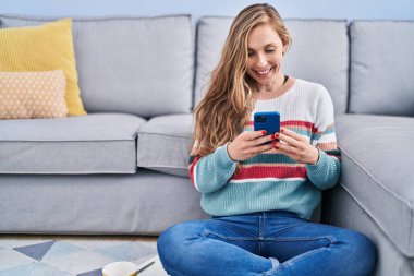 Young blonde woman using smartphone sitting on floor at home