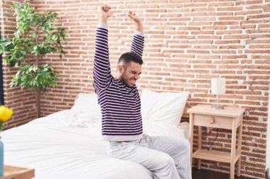 Young hispanic man waking up stretching arms at bedroom