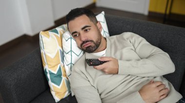 Evdeki sıkıcı ifadesiyle kanepede uzanmış televizyon izleyen İspanyol genç adam.
