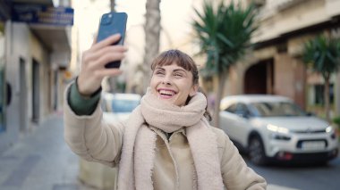 Beyaz bir kadın sokakta akıllı telefonuyla selfie çekiyor.