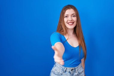 Mavi arka planda duran kızıl saçlı kadın selamlama ve hoş geldin demek için dostça el sıkışma teklif ediyor. başarılı iş. 