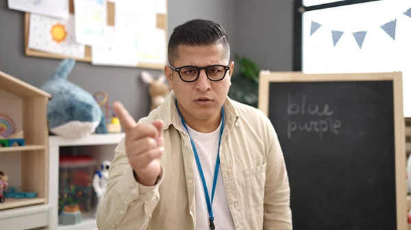 Young hispanic man preschool teacher with unhappy expression saying no with finger at kindergarten