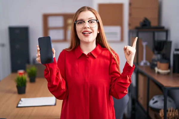 在办公室工作的红头发女人微笑着 手指手画脚地看着旁边的智能手机屏幕 — 图库照片