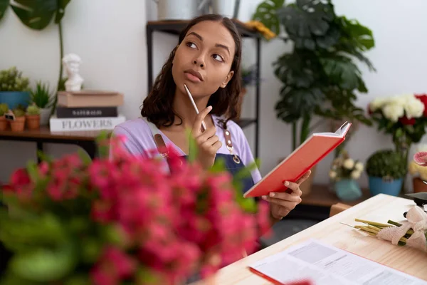 Young african american woman florist reading notebook with doubt expression at flower shop