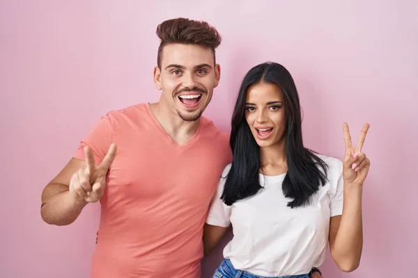 若いヒスパニック系のカップルがピンクの背景の上に立って幸せな顔が指で勝利のサインを行うカメラでウィンクと笑みを浮かべて — ストック写真