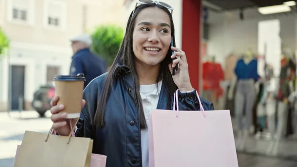 若い美しいヒスパニック系の女性が路上で買い物袋やコーヒーを持っているスマートフォンで話しています — ストック写真