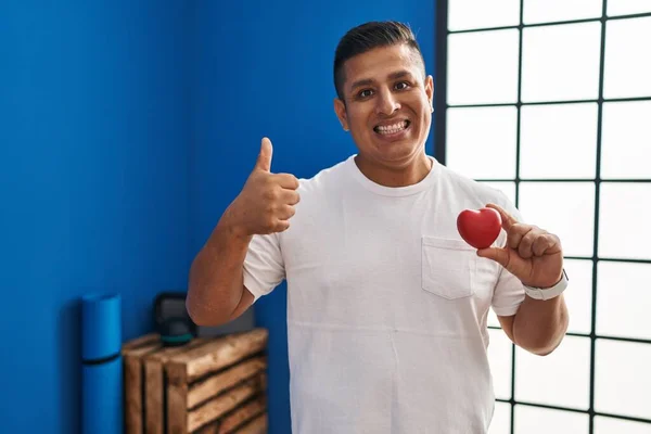 Hispanic young man holding red heart at gym smiling happy and positive, thumb up doing excellent and approval sign