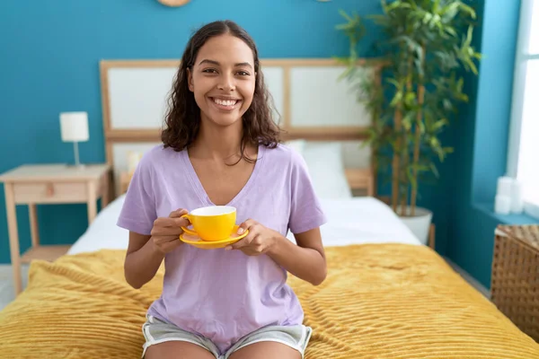 Young african american woman drinking cup of coffee sitting on bed at bedroom