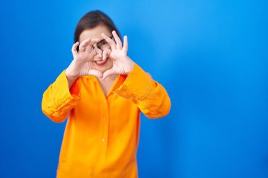 Mavi arka planda duran gözlüklü, orta yaşlı İspanyol bir kadın. Sevgiyle gülümseyen, elleriyle kalp sembolü yapan. romantik kavram. 