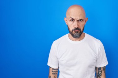 Mavi arka planda duran İspanyol dövmeli adam depresyonda ve üzüntüden ağlıyor, kızgın ve korkmuş. hüzünlü ifade. 