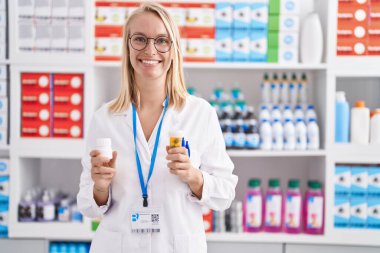 Young blonde woman pharmacist smiling confident holding pills bottles at pharmacy