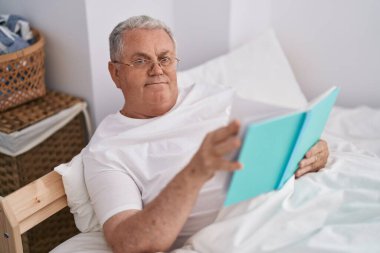 Orta yaşlı, kır saçlı, kitap okuyan, yatak odasında oturan bir adam.