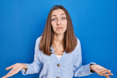 Young woman standing over blue background clueless and confused with open arms, no idea concept. 