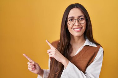 Sarı arka planda duran genç esmer kadın. Gözlüklü, gülümseyen ve kameraya bakan. İki eli ve parmağı yanda olan.. 