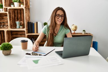 Genç İspanyol kadın dizüstü bilgisayar kullanıyor ve panoya yazı yazıyor. Evdeki masada oturuyor.