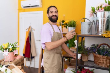 Young hispanic man florist putting plants on shelving at flower shop