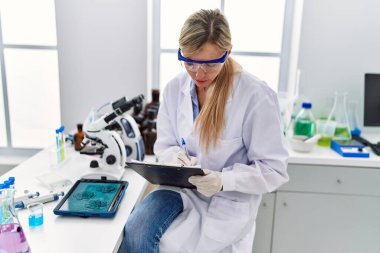 Young blonde woman wearing scientist uniform writing on clipboard looking embryion images at laboratory