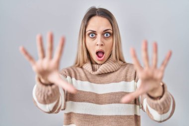 Sarışın bir kadın, izole edilmiş arka planda balıkçı yaka kazak giyiyor. Korkudan dehşete düşmüş ve korkmuş. El hareketlerini durdur, şok içinde bağır. panik konsepti. 