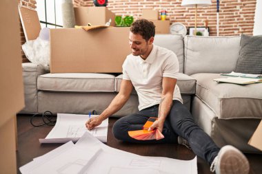 Young hispanic man writing on plans sitting on floor at new home