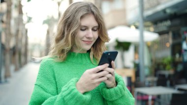 Young blonde woman smiling confident using smartphone at coffee shop terrace
