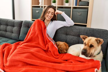 Young hispanic woman covering with blanket sitting on sofa with dogs at home