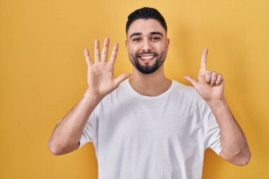 Sarı arka plan üzerine gündelik tişört giyen genç yakışıklı adam yedinci parmaklarını gösterip gülümserken kendinden emin ve mutlu görünüyor.. 