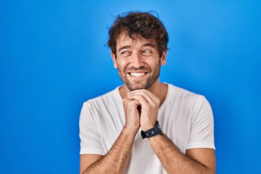 Mavi arka planda dikilen Latin asıllı genç adam. Gergin ve heyecanlı. Elleri çenesinde yan tarafa bakıyor. 