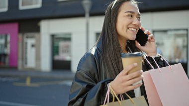 Güzel İspanyol bir kadın akıllı telefondan konuşuyor. Elinde alışveriş poşetleri ve sokakta kahve var.