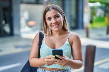 Young beautiful hispanic woman student smiling confident using smartphone at street