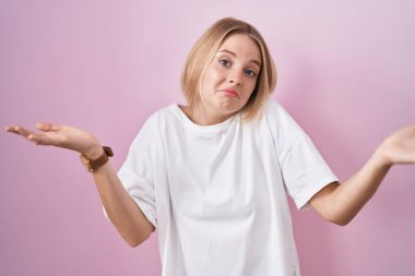 Pembe arka planda duran beyaz bir kadın. Kolları ve elleri havada, kafası karışmış bir ifade. şüphe kavramı. 