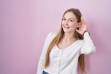 Young caucasian woman standing over pink background smiling with hand over ear listening an hearing to rumor or gossip. deafness concept. 