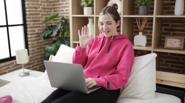 Young blonde woman having video call sitting on bed at bedroom