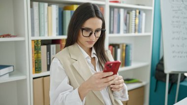 Genç, güzel İspanyol kız öğrenci üniversite sınıfında rahat bir ifadeyle akıllı telefon kullanıyor.