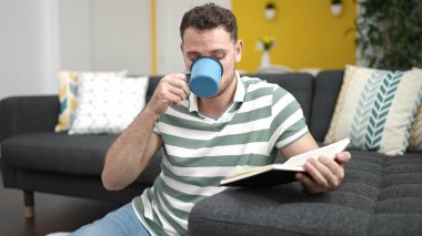 Genç beyaz adam kanepede oturmuş kitap okuyor. Evde kahve içiyor.