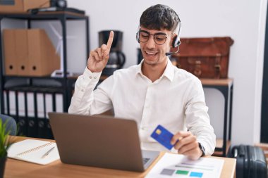 Bilgisayarda çalışan İspanyol genç adam elinde kredi kartıyla başarılı bir fikir işaret ediyor. Heyecanlı ve mutlu. Bir numara.. 