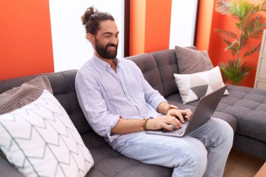 Young hispanic man using laptop sitting on sofa at home